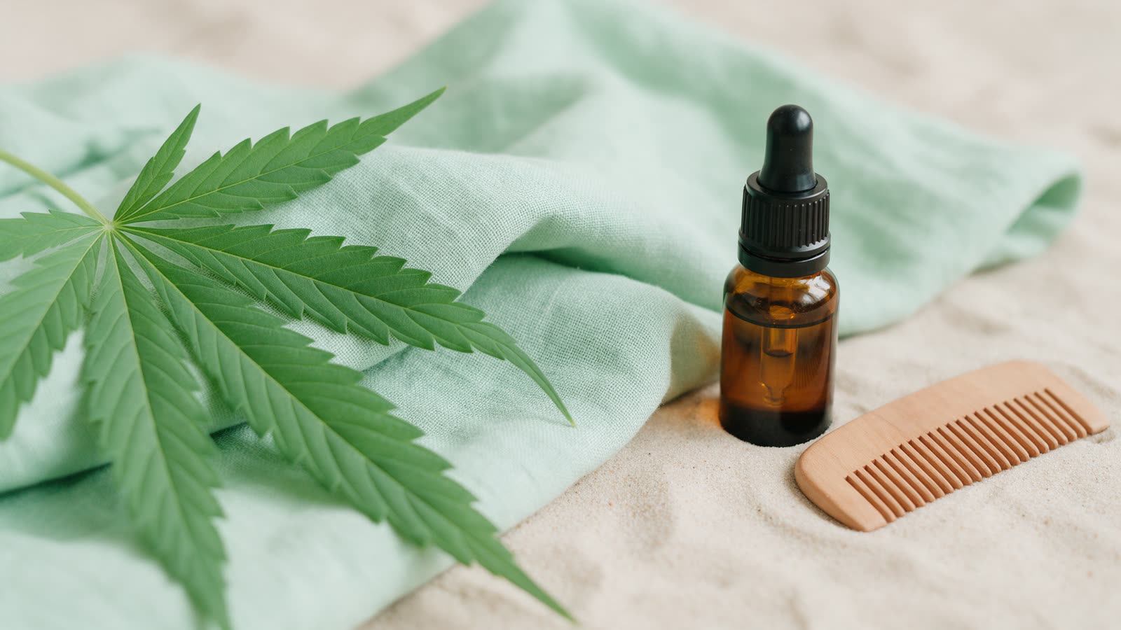 KinderHemp.com: gentle hemp wellness brand domain Fresh hemp leaves, folded hemp linen, an amber dropper bottle and a wooden comb on sand